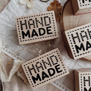 May include: Rectangular wooden stamps featuring the text HAND MADE in a bold black font. The letters are surrounded by a decorative border resembling cross-stitch embroidery. The stamps are made of light-colored wood with a smooth finish.