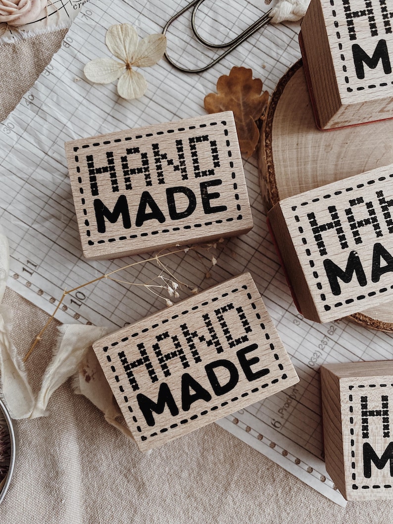 May include: Rectangular wooden stamps featuring the text HAND MADE in a bold black font. The letters are surrounded by a decorative border resembling cross-stitch embroidery. The stamps are made of light-colored wood with a smooth finish.
