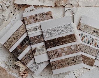Puede incluir: Una colección de rollos de cinta washi decorativa con varios diseños, incluyendo copos de nieve, diseños geométricos y escritura. Los rollos de cinta están dispuestos sobre un fondo de papel vintage, encaje y flores secas.