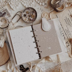 May include: A six-ring binder planner with a light beige magnetic closure tab. The open planner features two distinct types of paper: a textured off-white sheet with visible flecks and a smooth, speckled light brown sheet. The binder rings are metal.