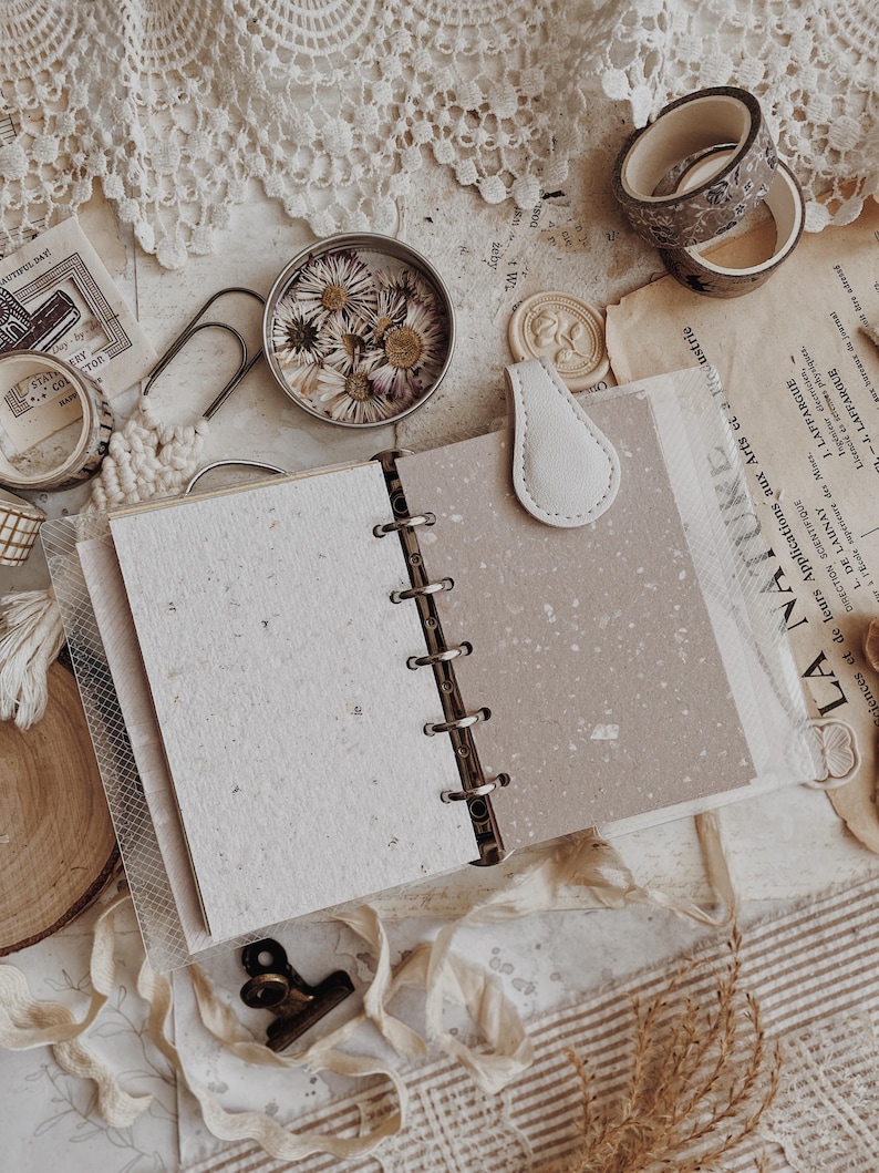 May include: A six-ring binder planner with a light beige magnetic closure tab. The open planner features two distinct types of paper: a textured off-white sheet with visible flecks and a smooth, speckled light brown sheet. The binder rings are metal.