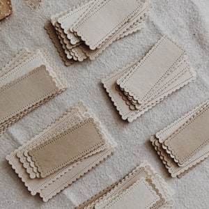 Puede incluir: Varias pilas de etiquetas rectangulares de papel en tonos beige y marr&oacute;n claro se disponen sobre una superficie de lino neutra. Cada etiqueta tiene un borde festoneado y un borde de l&iacute;nea punteada. Ideales para manualidades.