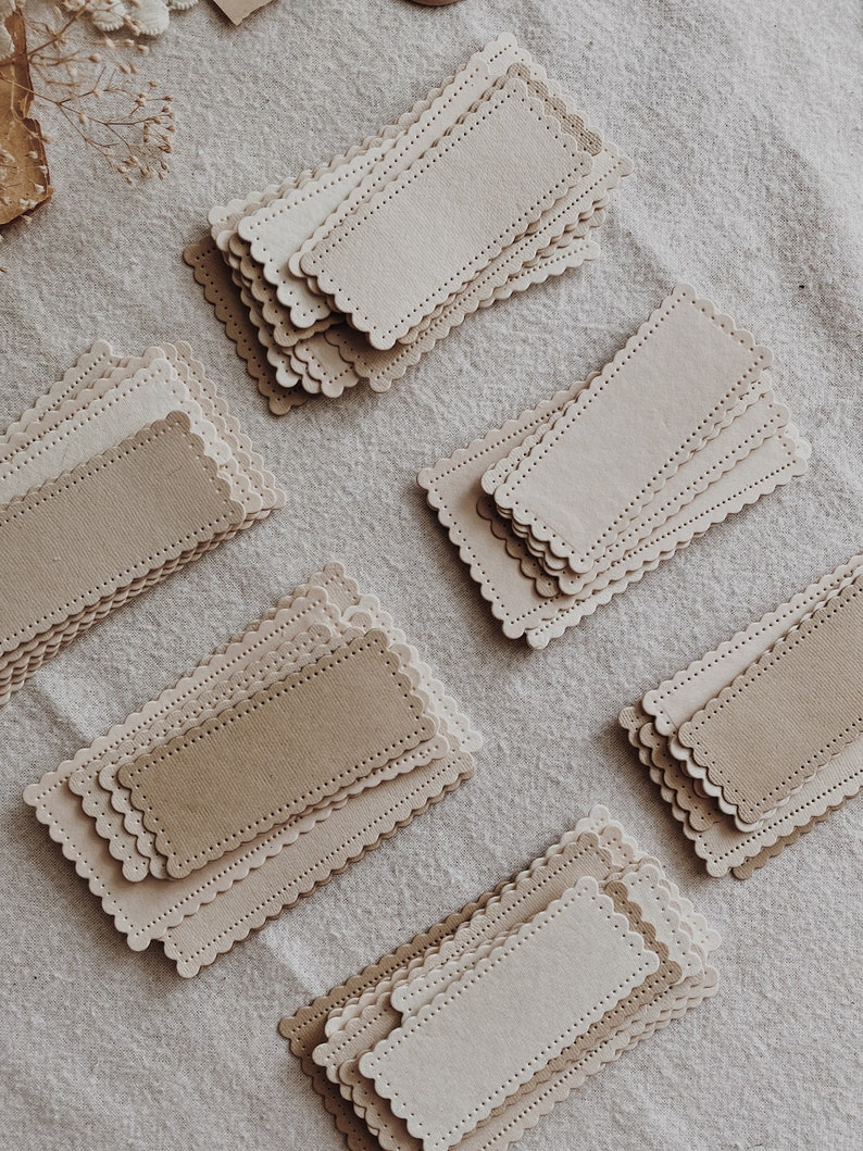 Puede incluir: Varias pilas de etiquetas rectangulares de papel en tonos beige y marr&oacute;n claro se disponen sobre una superficie de lino neutra. Cada etiqueta tiene un borde festoneado y un borde de l&iacute;nea punteada. Ideales para manualidades.