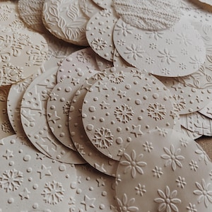 May include: A collection of off-white, circular paper ornaments with embossed patterns. Designs include snowflakes, stars, flowers, and textured knit-like patterns. The ornaments are scattered, creating a layered effect.