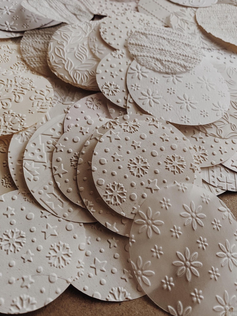 May include: A collection of off-white, circular paper ornaments with embossed patterns. Designs include snowflakes, stars, flowers, and textured knit-like patterns. The ornaments are scattered, creating a layered effect.