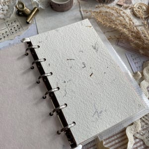 May include: A ring-bound notebook featuring off-white, textured paper with visible organic flecks. The binding consists of a silver-toned metal spine with circular rings holding the pages securely in place, providing a functional and simple design.