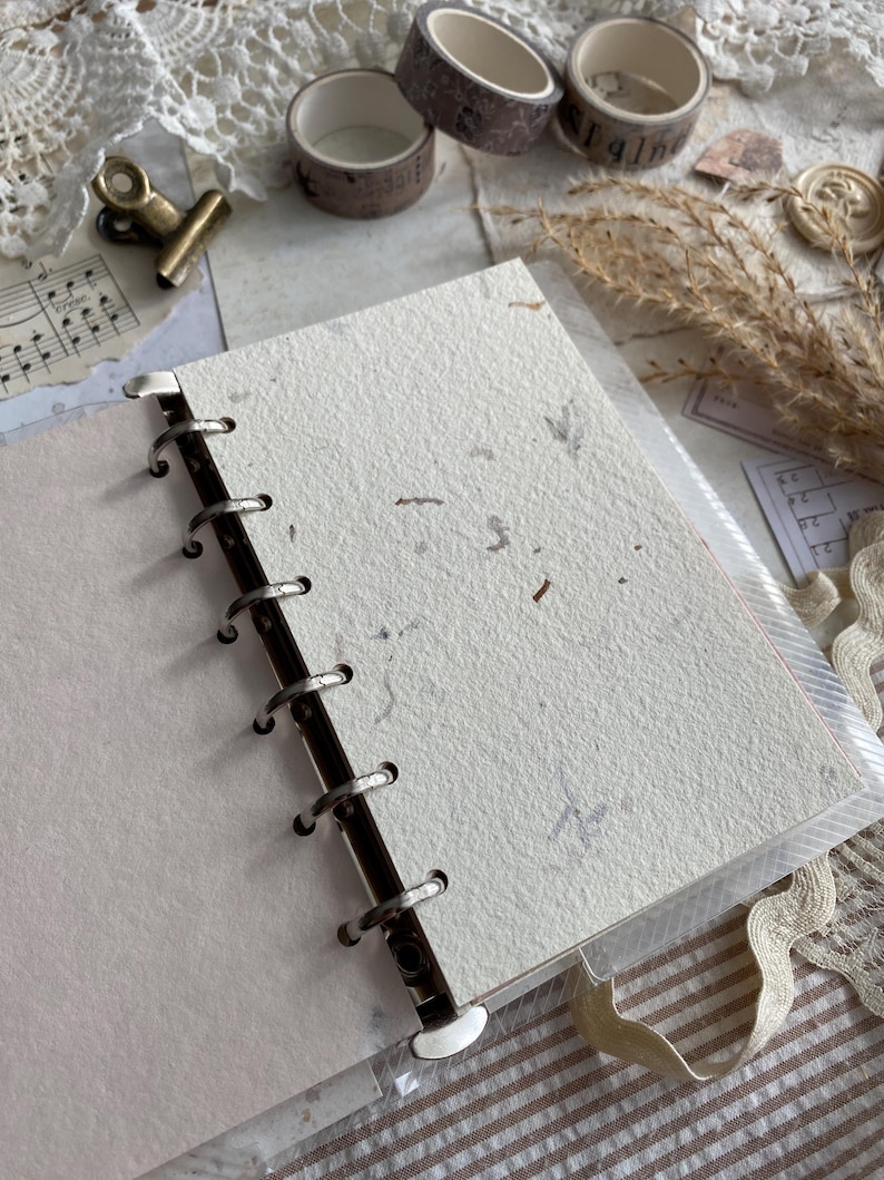 May include: A ring-bound notebook featuring off-white, textured paper with visible organic flecks. The binding consists of a silver-toned metal spine with circular rings holding the pages securely in place, providing a functional and simple design.