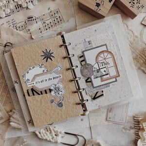 May include: A ring-bound journal with tan and speckled white pages featuring various paper cutouts, including a star, a flower, and a rectangular label with the text 'it's all in the details', plus a window graphic with the text 'This.'