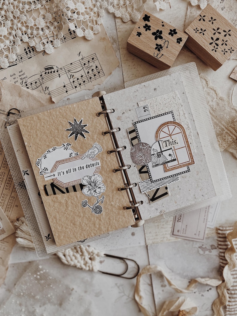 May include: A ring-bound journal with tan and speckled white pages featuring various paper cutouts, including a star, a flower, and a rectangular label with the text 'it's all in the details', plus a window graphic with the text 'This.'