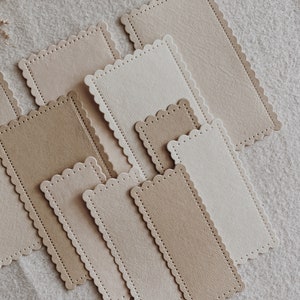 Puede incluir: Una colecci&oacute;n de etiquetas rectangulares de papel con bordes festoneados en varios tonos de beige y marr&oacute;n. Algunas etiquetas tienen una superficie texturizada, otras son lisas. Las etiquetas est&aacute;n dispuestas sobre un fondo de tela de color neutro.