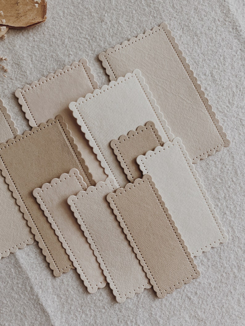 Puede incluir: Una colecci&oacute;n de etiquetas rectangulares de papel con bordes festoneados en varios tonos de beige y marr&oacute;n. Algunas etiquetas tienen una superficie texturizada, otras son lisas. Las etiquetas est&aacute;n dispuestas sobre un fondo de tela de color neutro.
