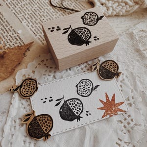 May include: A rectangular wooden rubber stamp featuring a black ink pomegranate design. Below the stamp, a white rectangular card displays the same black pomegranate print alongside a small, textured, rust-colored star shape.