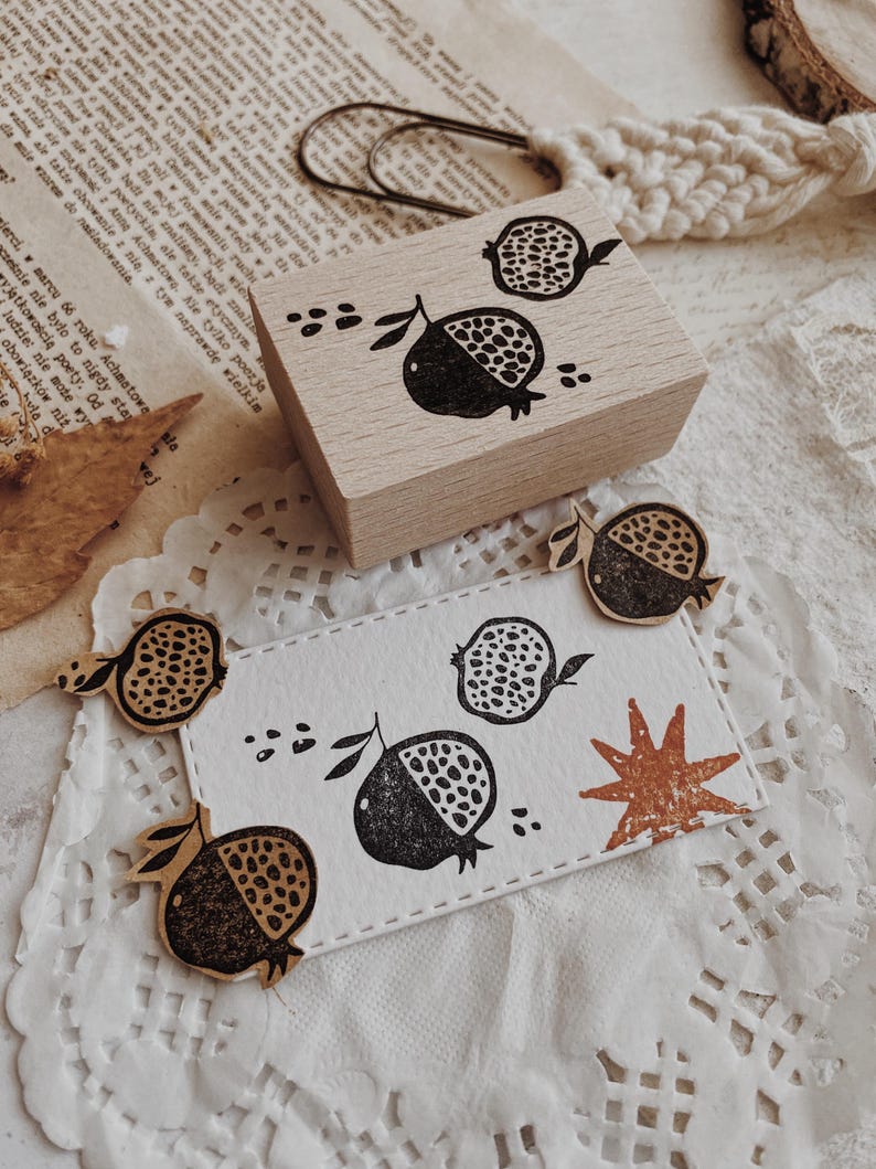 May include: A rectangular wooden rubber stamp featuring a black ink pomegranate design. Below the stamp, a white rectangular card displays the same black pomegranate print alongside a small, textured, rust-colored star shape.