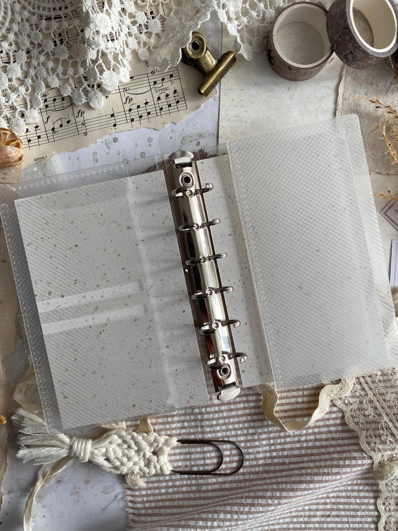 May include: A clear plastic six-ring binder with a silver metal mechanism. The binder features translucent covers with a subtle diagonal grid texture and contains off-white paper inserts with small dark flecks throughout the pages.