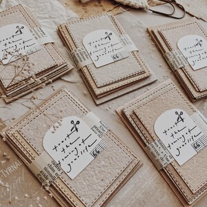 May include: Stacks of beige paper cards with stitched edges are bundled together. Each bundle is wrapped with a strip of paper and a white tag that reads "trust the framing of your life." The cards have a rustic, handmade aesthetic.