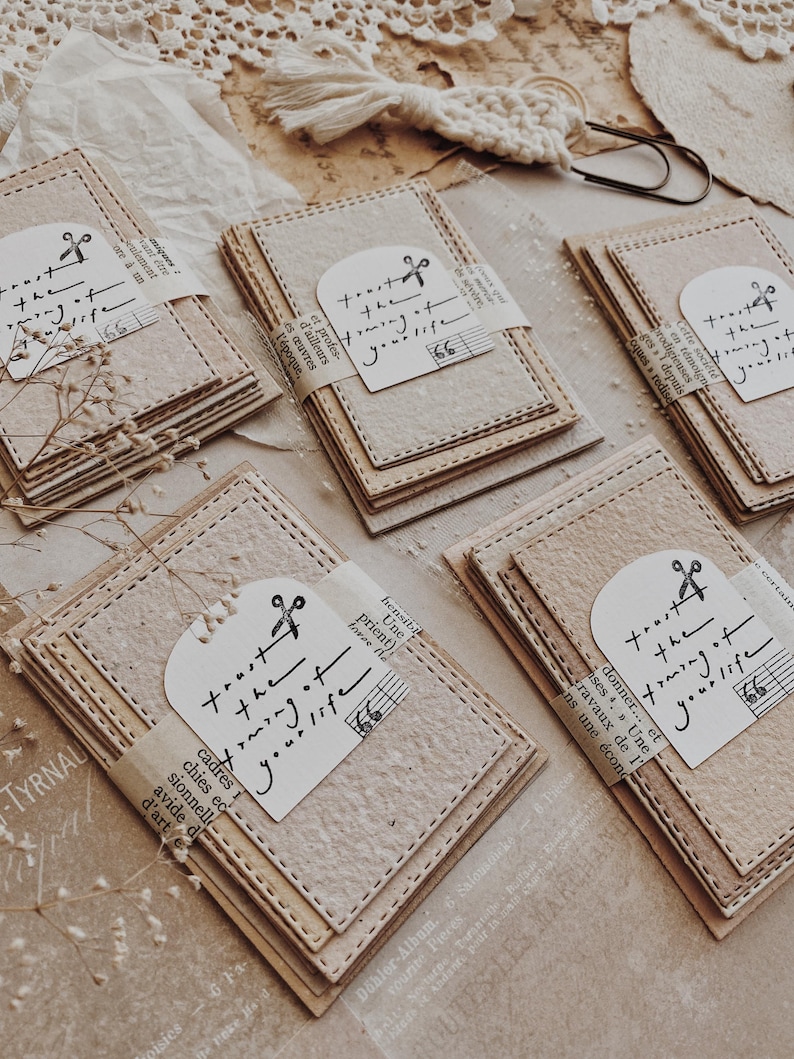 May include: Stacks of beige paper cards with stitched edges are bundled together. Each bundle is wrapped with a strip of paper and a white tag that reads "trust the framing of your life." The cards have a rustic, handmade aesthetic.