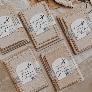 May include: A collection of beige paper notecards with stitched edges, each wrapped with a paper band. The cards feature a printed design with the text "trust the timing of your life" and a musical note.