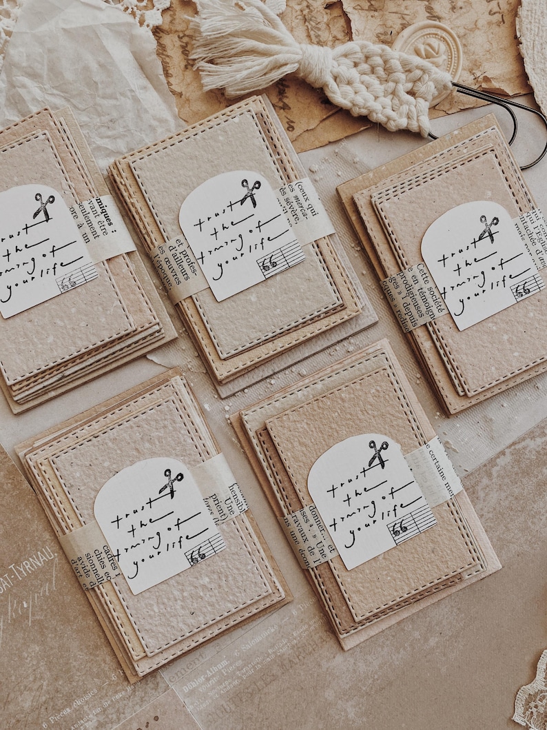 May include: A collection of beige paper notecards with stitched edges, each wrapped with a paper band. The cards feature a printed design with the text "trust the timing of your life" and a musical note.