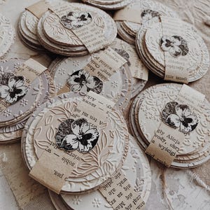 May include: A collection of handmade, round paper crafts with embossed floral designs and attached paper tags. Each craft features a black and white floral image. The crafts are in varying shades of cream and beige.