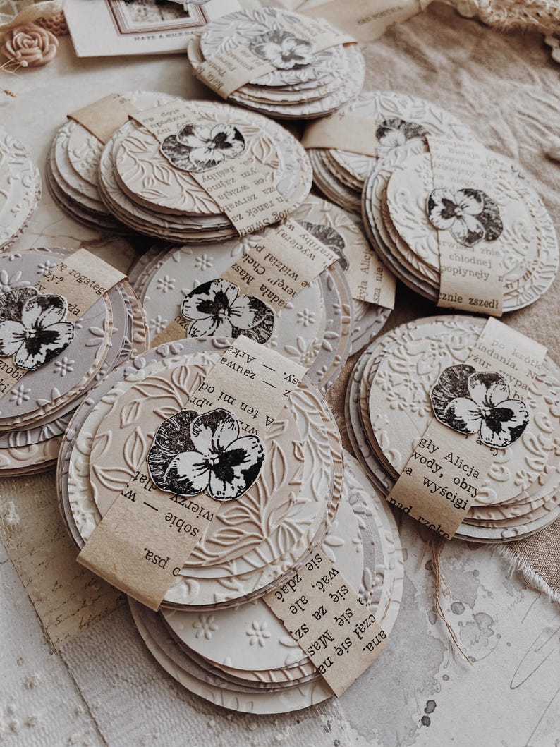 May include: A collection of handmade, round paper crafts with embossed floral designs and attached paper tags. Each craft features a black and white floral image. The crafts are in varying shades of cream and beige.