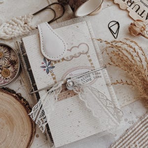 Puede incluir: Un diario con cierre de lengüeta de cuero blanco, adornado con encaje, un broche de metal y un dije de concha. Cinta washi decorativa, clips y flores secas rodean el diario, creando una estética vintage.