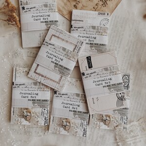 May include: A set of six vintage-style journaling cards with a distressed paper texture. Each card features the text "Journaling Card Set" and "Here We Are!" in a typewriter font. The cards are decorated with various vintage ephemera, including stamps, labels, and paper scraps.