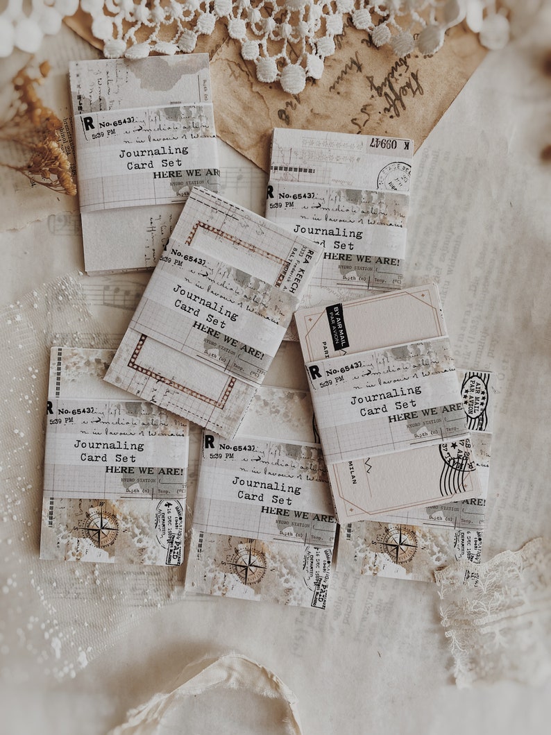 May include: A set of six vintage-style journaling cards with a distressed paper texture. Each card features the text "Journaling Card Set" and "Here We Are!" in a typewriter font. The cards are decorated with various vintage ephemera, including stamps, labels, and paper scraps.