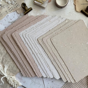 May include: A stack of rectangular handmade paper sheets with deckled edges in varying shades of beige, cream, and light brown. The paper has a visible fibrous texture with small flecks throughout, arranged in a fan shape on a flat surface.