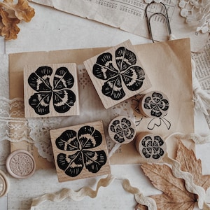 Puede incluir: Sellos de madera con un diseño de trébol de cuatro hojas. Los sellos son de varios tamaños y formas, con un patrón de trébol negro detallado. La imagen también incluye elementos decorativos como encaje, cintas y hojas secas.
