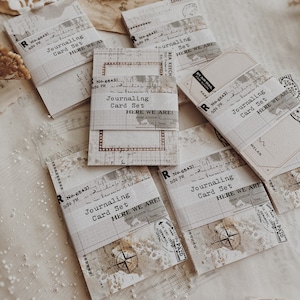 May include: Five vintage-style journaling card sets with a distressed white background and black text. Each card set has a label that reads "Journaling Card Set" and "Here We Are!" with a vintage stamp and a compass design.