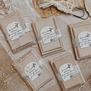 May include: A collection of handmade paper cards with stitched edges, tied with a paper band featuring the text "trust the forming of your life" and a scissor graphic. The cards are beige and cream colored.