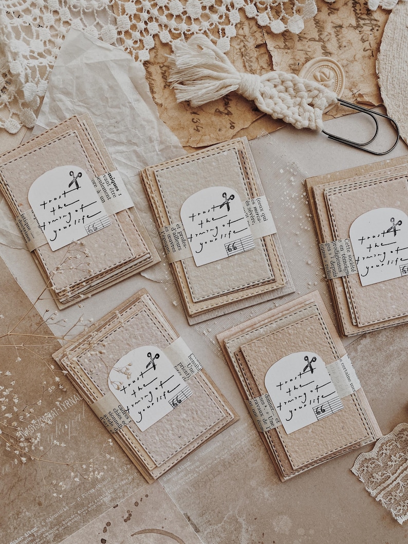 May include: A collection of handmade paper cards with stitched edges, tied with a paper band featuring the text "trust the forming of your life" and a scissor graphic. The cards are beige and cream colored.