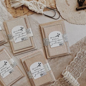 May include: A collection of handmade paper tags with the text "trust the timing of your life" and musical notes. The tags are tied with a strip of paper and are surrounded by vintage-style lace and paper.