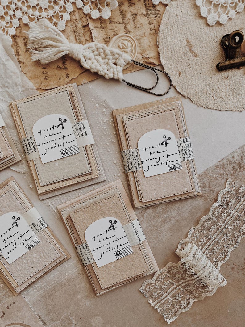 May include: A collection of handmade paper tags with the text "trust the timing of your life" and musical notes. The tags are tied with a strip of paper and are surrounded by vintage-style lace and paper.