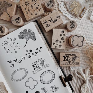 Op de afbeelding: Een verzameling houten rubberen stempels met diverse ontwerpen zoals sterren, vogels, bladeren, paddenstoelen en teksten zoals Parcel Post No 4, gerangschikt naast een notitieboek met zwarte inkt-afdrukken van elk ontwerp.