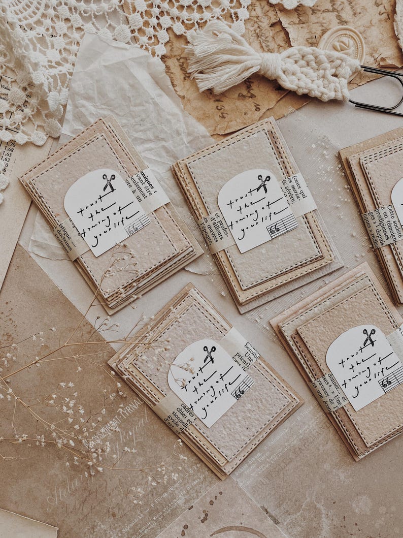 May include: A collection of handmade paper cards with stitched edges, each wrapped with a paper band and a label that reads "trust the timing of your life." The cards are arranged on a neutral-toned surface.