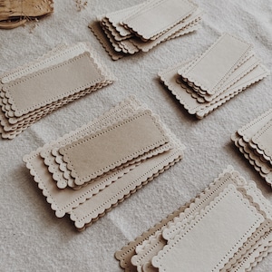 Puede incluir: Varias pilas de etiquetas rectangulares de papel, con bordes festoneados, dispuestas sobre una superficie de lino neutro. Las etiquetas son en tonos beige y crema. Cada etiqueta tiene un borde perforado y un &aacute;rea central ligeramente texturizada.