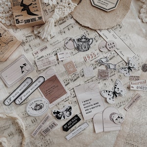 May include: A collection of vintage-style stickers and ephemera featuring butterflies, teapots, and other whimsical designs. The stickers are printed on white paper with black ink and have a distressed, aged look. The ephemera includes a small piece of music sheet, a vintage postcard, and a small piece of lace.