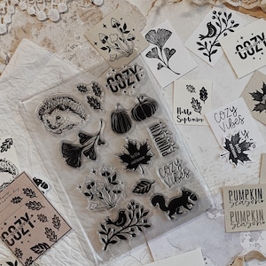 Könnte beinhalten: Ein transparenter Silikonstempelbogen mit herbstlichen Motiven wie einem Igel, Kürbissen, Pilzen, Blättern, einem Eichhörnchen und Vögeln. Der Text umfasst "COZY", "COZY VIBES", "PUMPKIN SEASON", "hello autumn" und "Hello September".