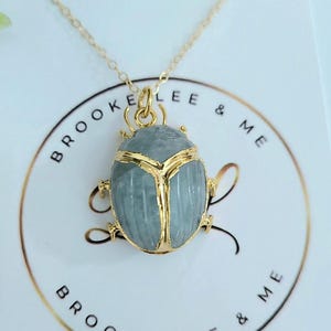 May include: A gold chain necklace with a blue scarab beetle pendant. The pendant is made of a light blue stone with gold accents. The necklace is displayed on a white card with the text "Brooke Lee & Me" printed on it.