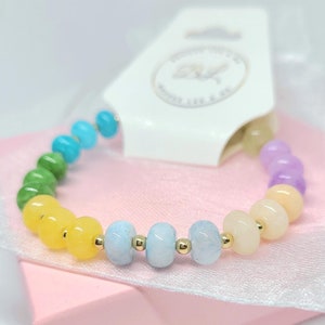 May include: A rainbow-colored beaded bracelet with gold spacers. The beads are in shades of purple, light pink, yellow, light blue, green, and turquoise.