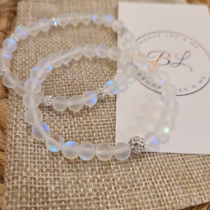 May include: Two iridescent white beaded bracelets with silver accents. The bracelets are stacked on top of each other and are on a burlap surface.