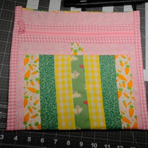 May include: A pink quilted zippered pouch with a patchwork design. The pouch features a pink gingham border, a pink zipper, and a central panel with carrot, bunny, and gingham patterns in green, yellow, and white.