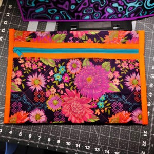 May include: A quilted fabric pouch with a vibrant floral pattern in pink, purple, and orange. The pouch has a teal zipper and orange trim. The background is a dark purple color.