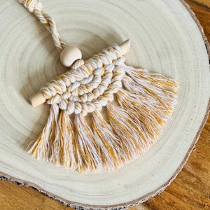 May include: A macrame wall hanging with a wooden dowel and a wooden bead. The macrame is made with white and mustard yellow yarn and has long fringe.