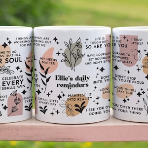 Puede incluir: Tres tazas de cer&aacute;mica blanca con dise&ntilde;os florales negros y rosas y citas inspiradoras. Las tazas presentan citas como "Disfruta de las cosas m&aacute;s peque&ntilde;as", "Celebra cada victoria", "Todo lo que necesito est&aacute; dentro de m&iacute;", y "Estoy agradecido".
