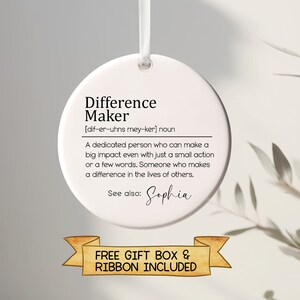 May include: White round bauble with the text "Difference Maker" and a definition of the word. The text "See also: Sophia" is below the definition. The bauble is hanging from a white ribbon. The text "FREE GIFT BOX & RIBBON INCLUDED" is below the bauble.