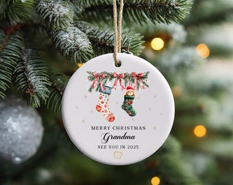 Op de afbeelding: Witte kerstbal van keramiek met een rood-groene krans, twee kousen en de tekst "Merry Christmas Oma Tot ziens in 2025".