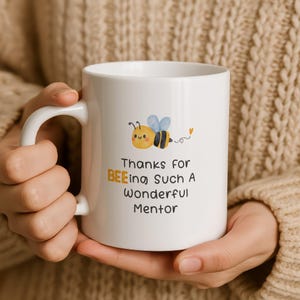 May include: A white ceramic mug featuring a cartoon bee illustration with blue wings and a yellow body. The text reads: Thanks for BEEing Such A Wonderful Mentor, with the word BEEing highlighted in yellow. The mug has a simple handle.