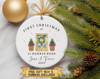 Op de afbeelding: Ronde witte kerstbal met een aquarel-illustratie van een huis met een krans op de deur, twee kleine kerstboompjes en de tekst "FIRST CHRISTMAS AT 21 HARRIS ROAD Jean & Trevor 2024".